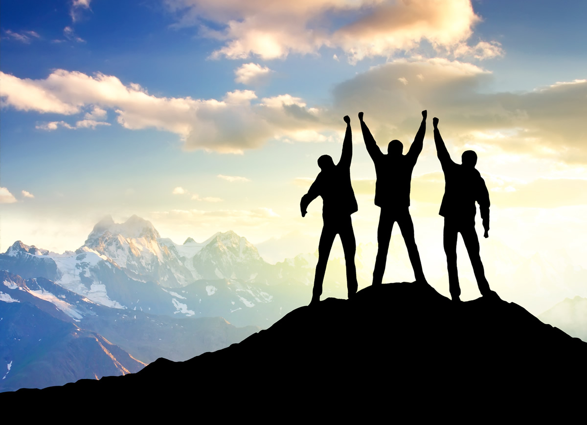Image of three business people silhouettes on a mountain top with arms raised in victory.